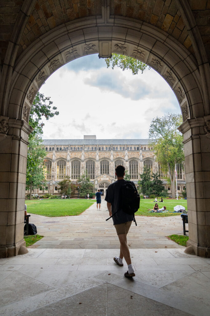 A student wearing shorts, a t-shirt, sandals, and a backpack walks through the arched stone walkway into the Law Quad.